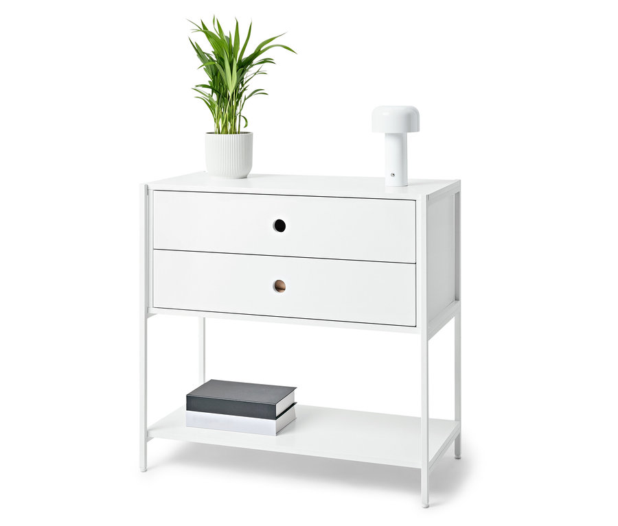 Biely nočný stolík s dvomi zásuvkami, lampou, rastlinou v kvetináči a knihami.