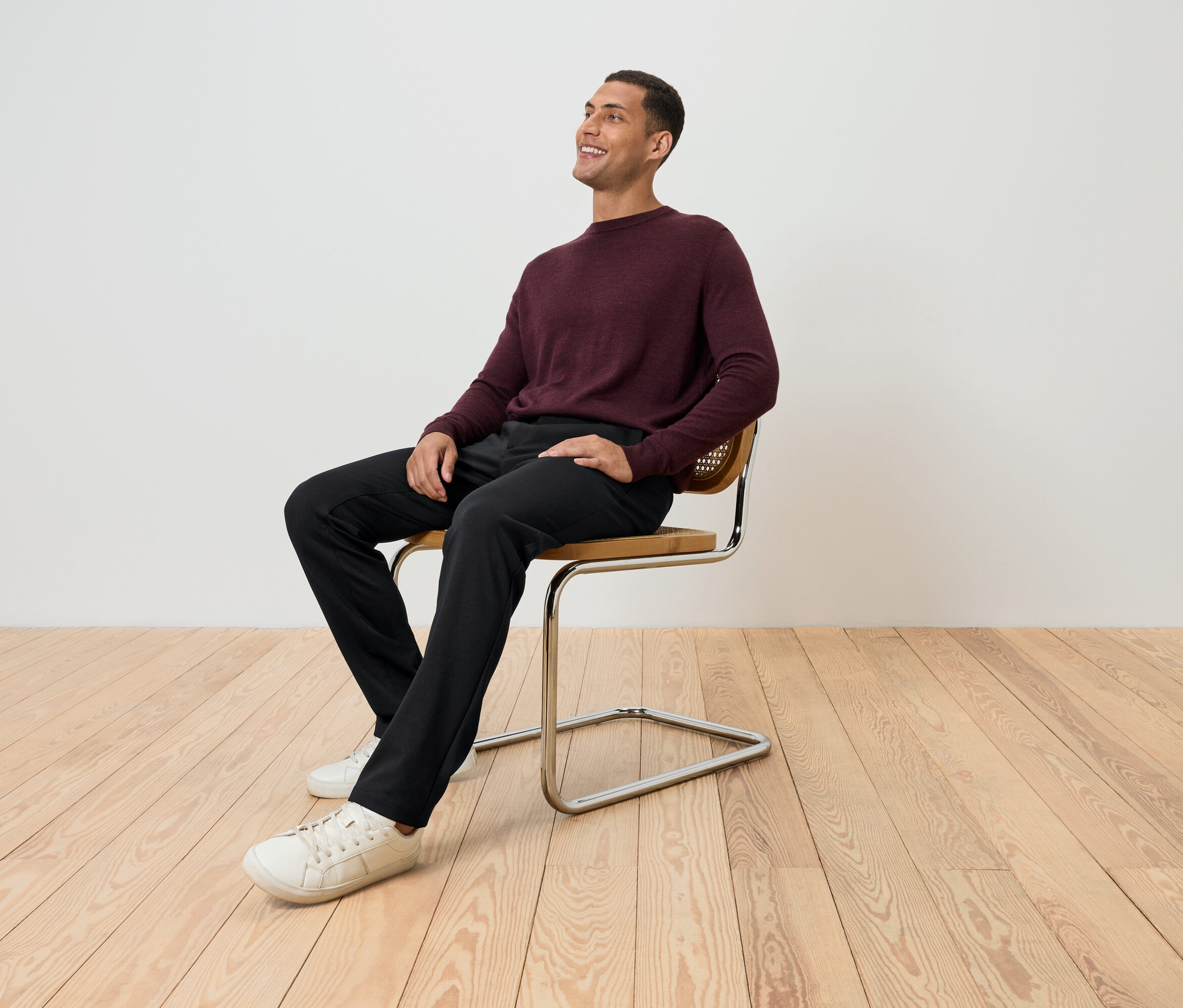 Muž sedí na stoličke v bordovom svetri, čiernych nohaviciach a bielych topánkach a pozerá sa hore.