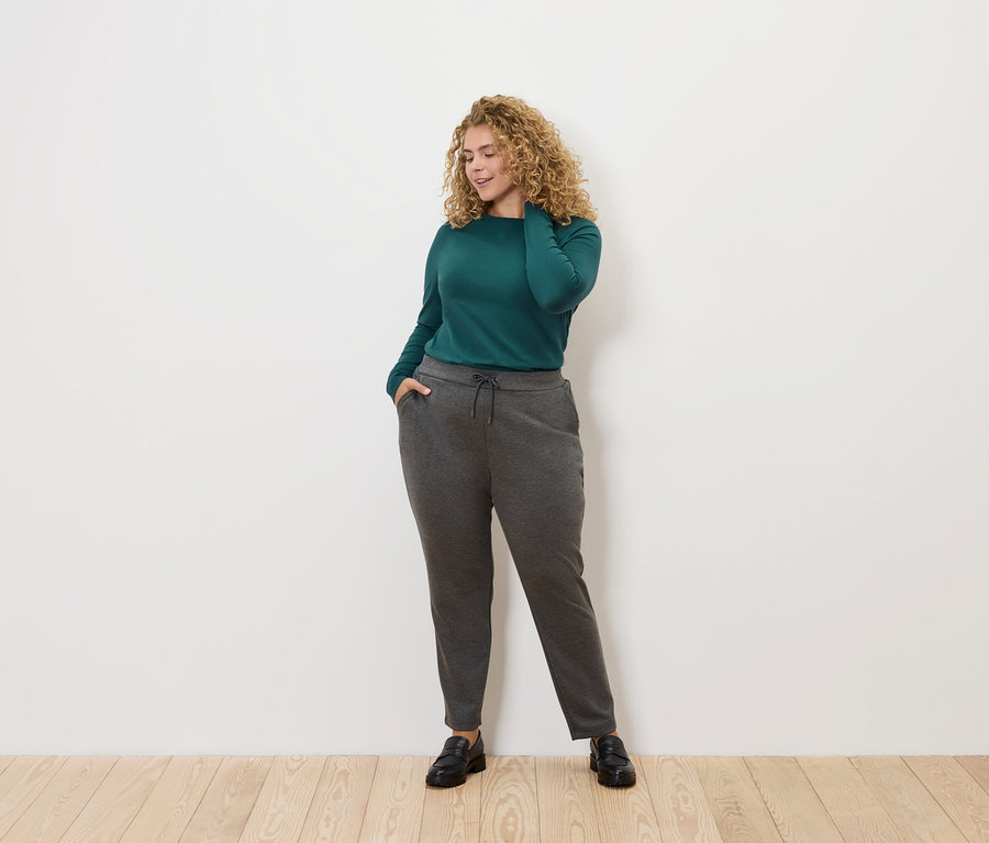 Žena s kučeravými vlasmi pózuje na svetlom pozadí. Má na sebe zelenú blúzku s dlhým rukávom a sivé tepláky.
