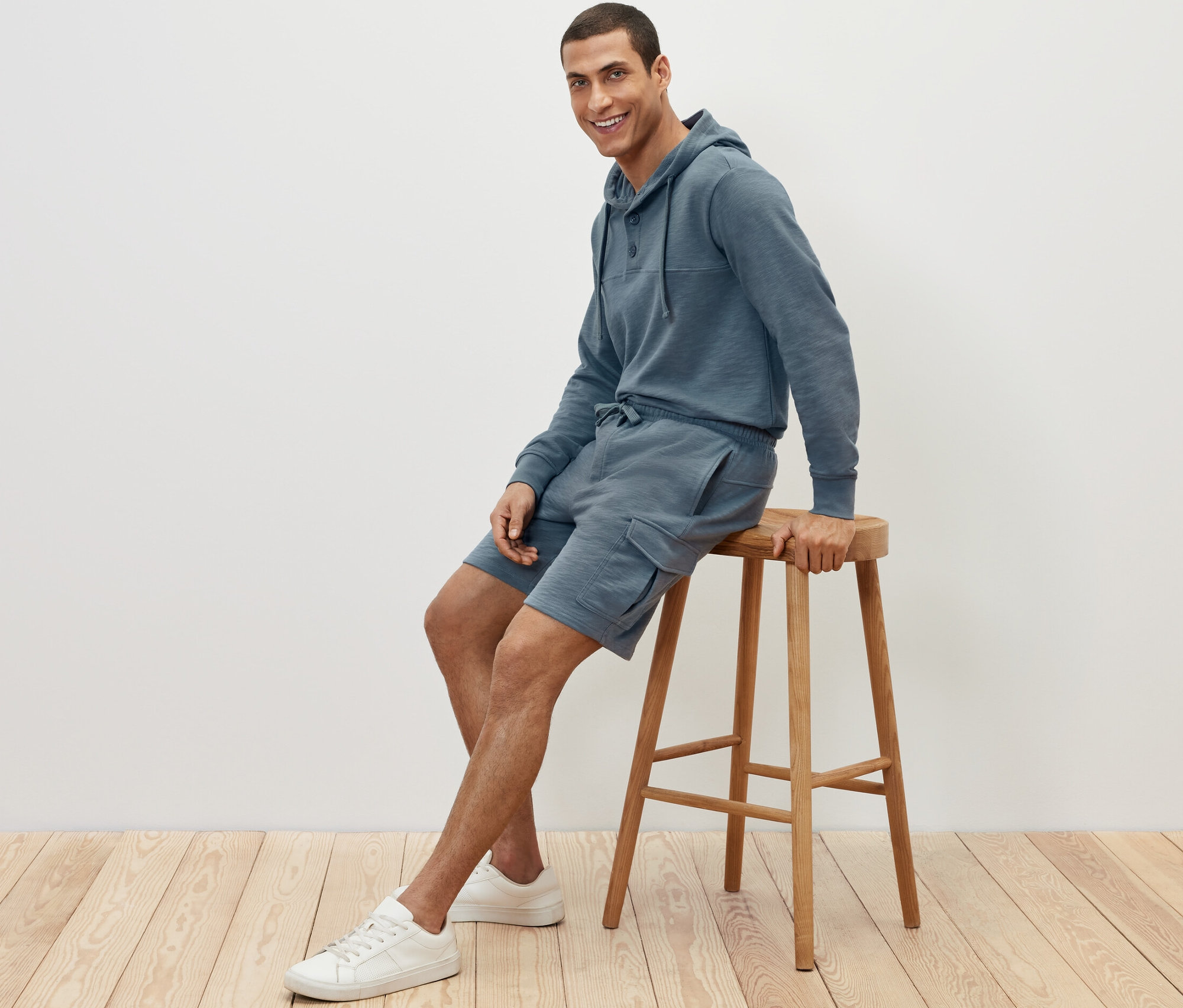 Muž sedí na stoličke a má na sebe cargo teplákové šortky a mikinu s kapucňou.