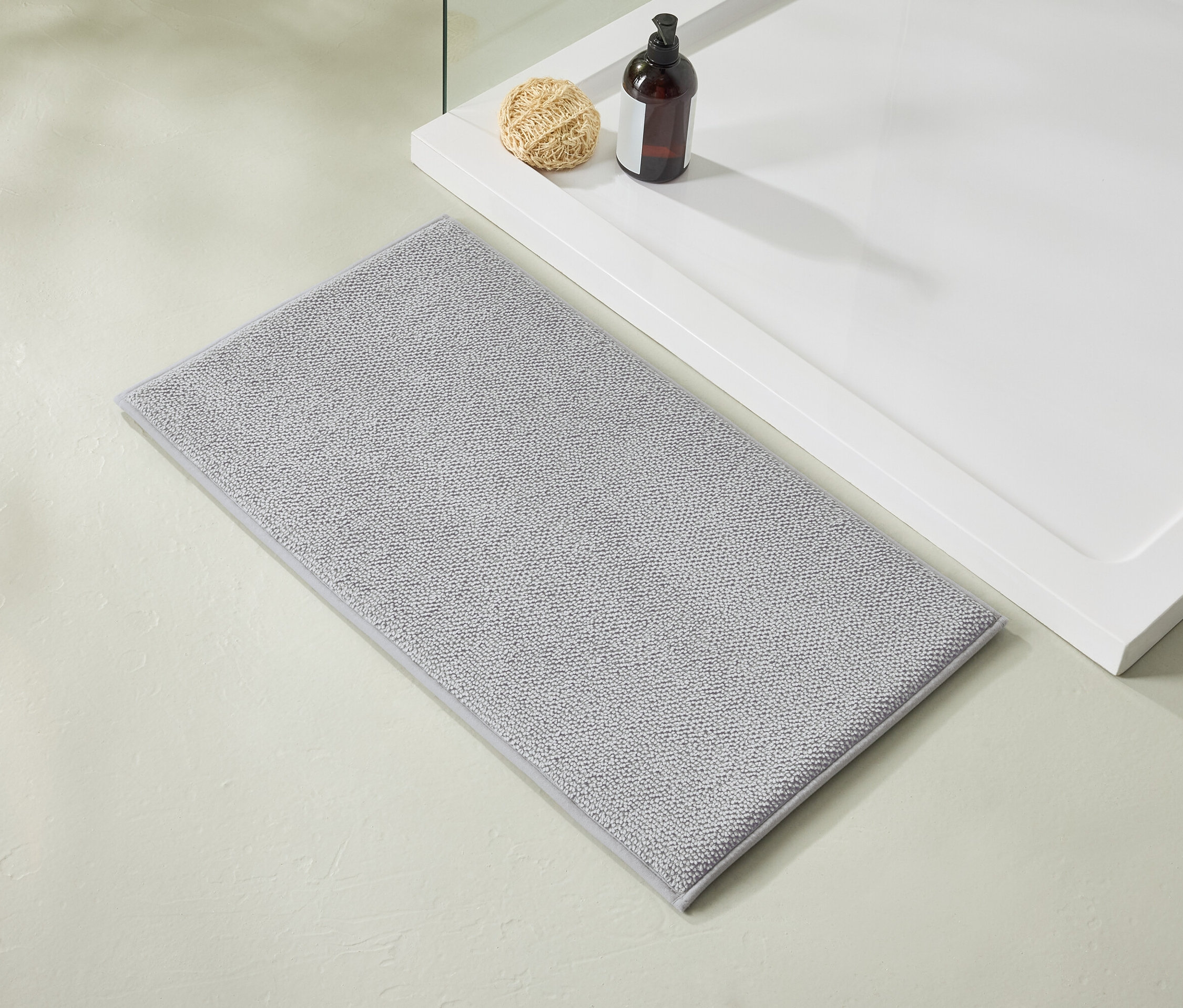 Svetlosivý kúpeľňový koberček leží vedľa bielej sprchovej vaničky, na ktorej stojí hnedá fľaštička a lufa.