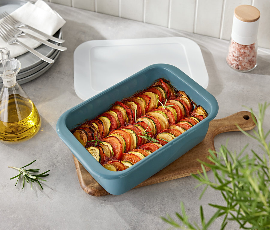 Zelenina v modrej zapekacej miske na drevenej doske. Vedľa misy je fľaša s olejom a koreničky.