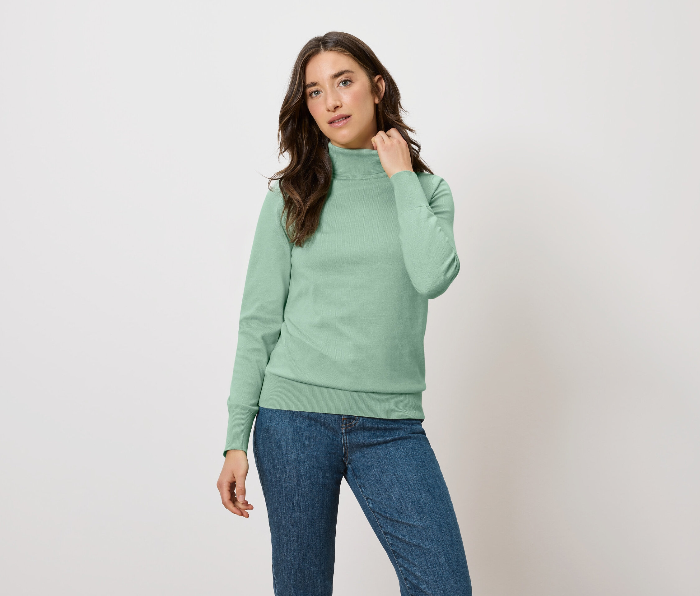 Žena pózuje v zelenej mikine s rolákom a modrých džínsoch.