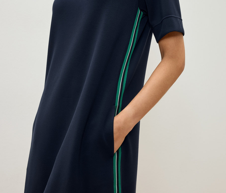 Žena má ruku vo vrecku tmavomodrých šiat s krátkym rukávom a zeleným a bielym pruhom.