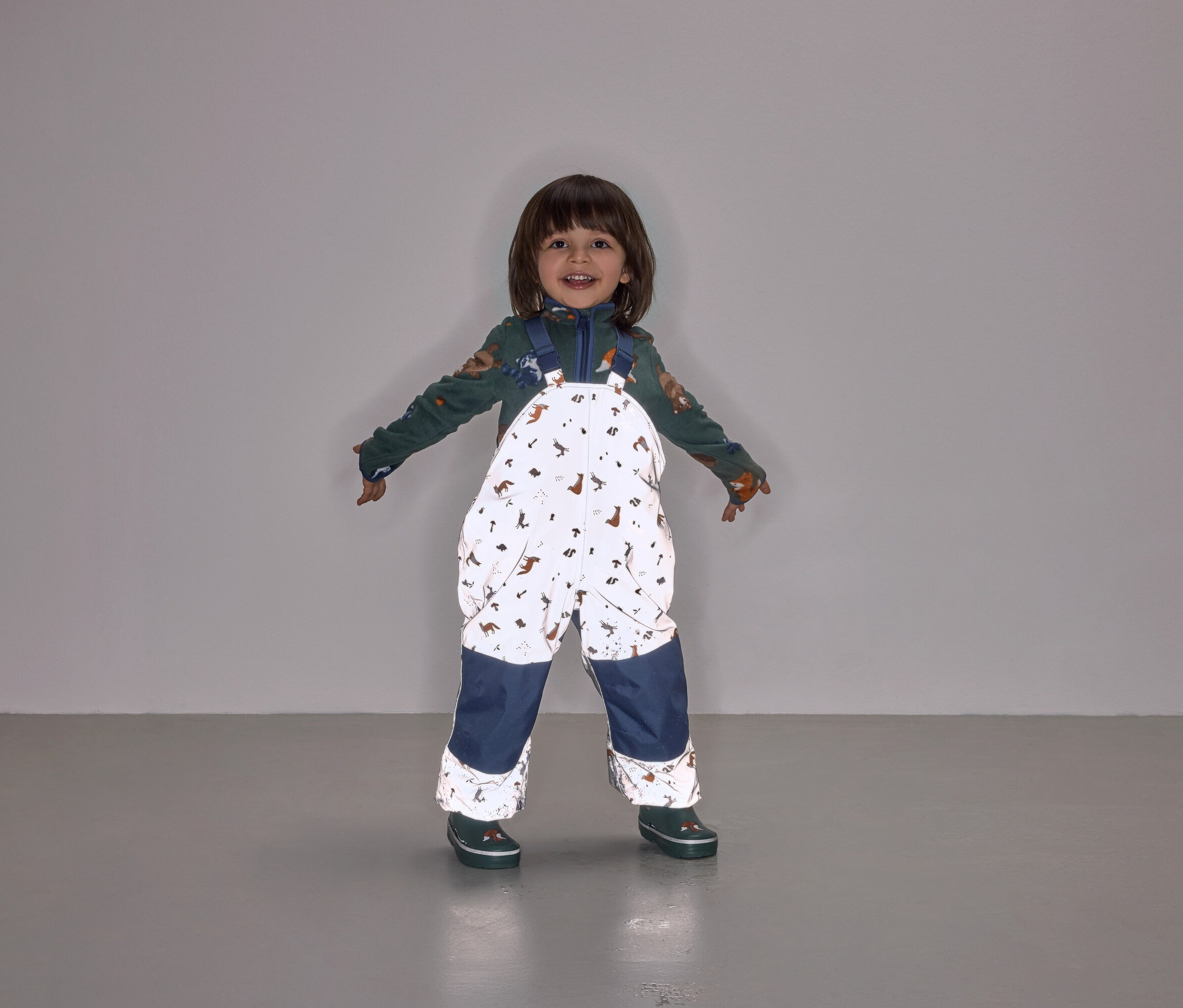Usmievajúce sa dieťa stojí s rozpaženými rukami a má na sebe kombinézu s náprsenkou s potlačou zvierat a tmavomodrými nohavicami.