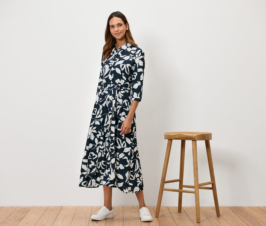 Žena stojí vedľa drevenej stoličky a má na sebe tmavomodré šaty s bielym kvetinovým vzorom.