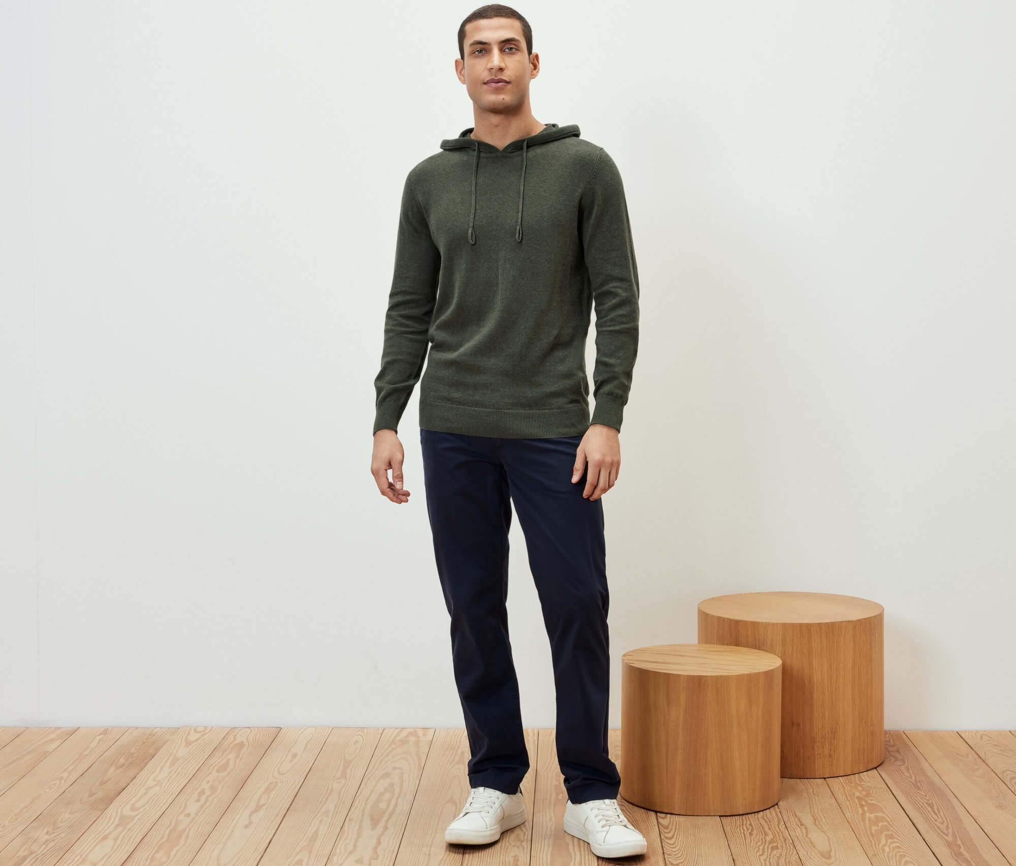 Muž stojí a má na sebe pletenú mikinu s kapucňou, tmavomodré nohavice a biele tenisky.