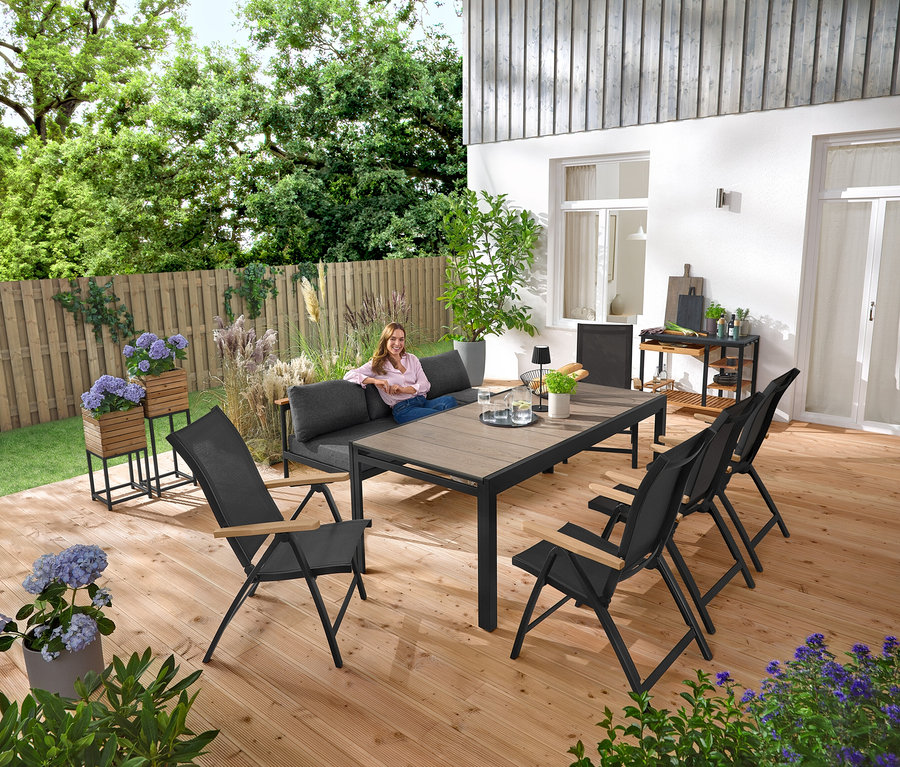 Žena sedí na záhradnej lavici 2 v 1 ‒ ležadle »Elin«. V pozadí sú 2 stĺpy na rastliny »Elin«. Na terase je tiež grilovací vozík s polycementovou pracovnou plochou a rozkladací záhradný stôl so stolovou doskou Duraboard.