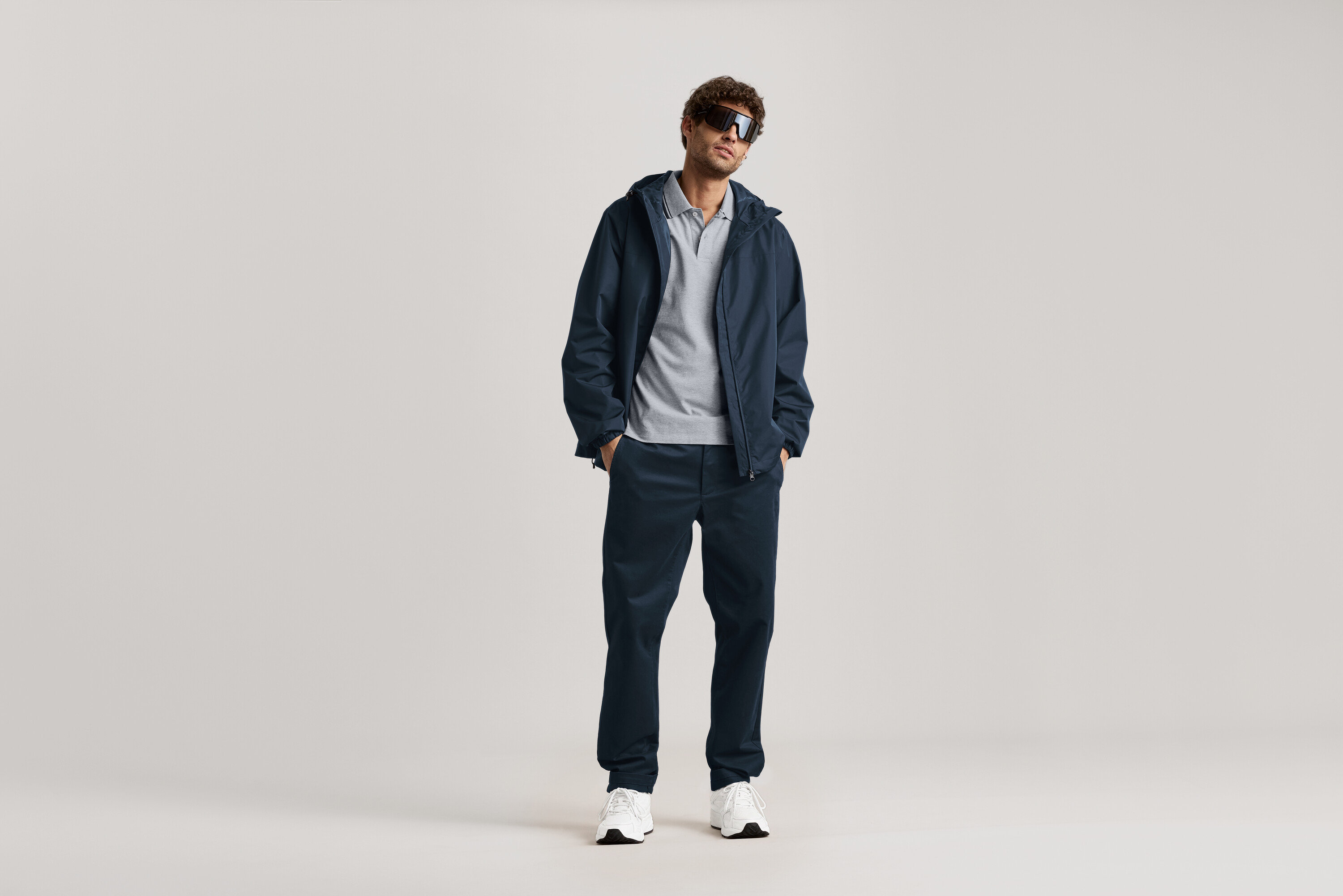Muž stojí so slnečnými okuliarmi, tmavomodrou bundou a tmavomodrými chino nohavicami.