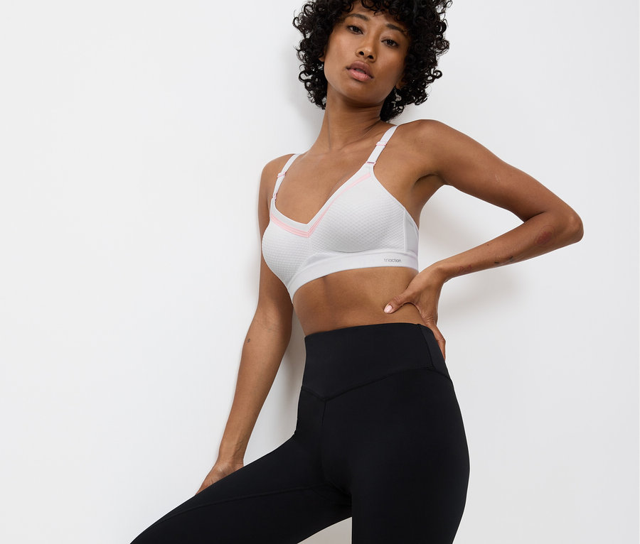 Žena pózuje v bielej športovej podprsenke s ružovými detailmi a čiernych legínach pred bielou stenou.