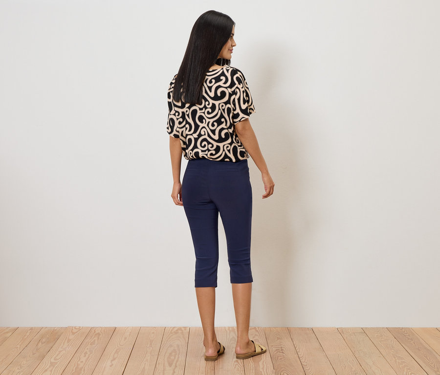 Žena zozadu v vzorovanej blúzke, modrých capri nohaviciach a sandáloch pred bielou stenou.