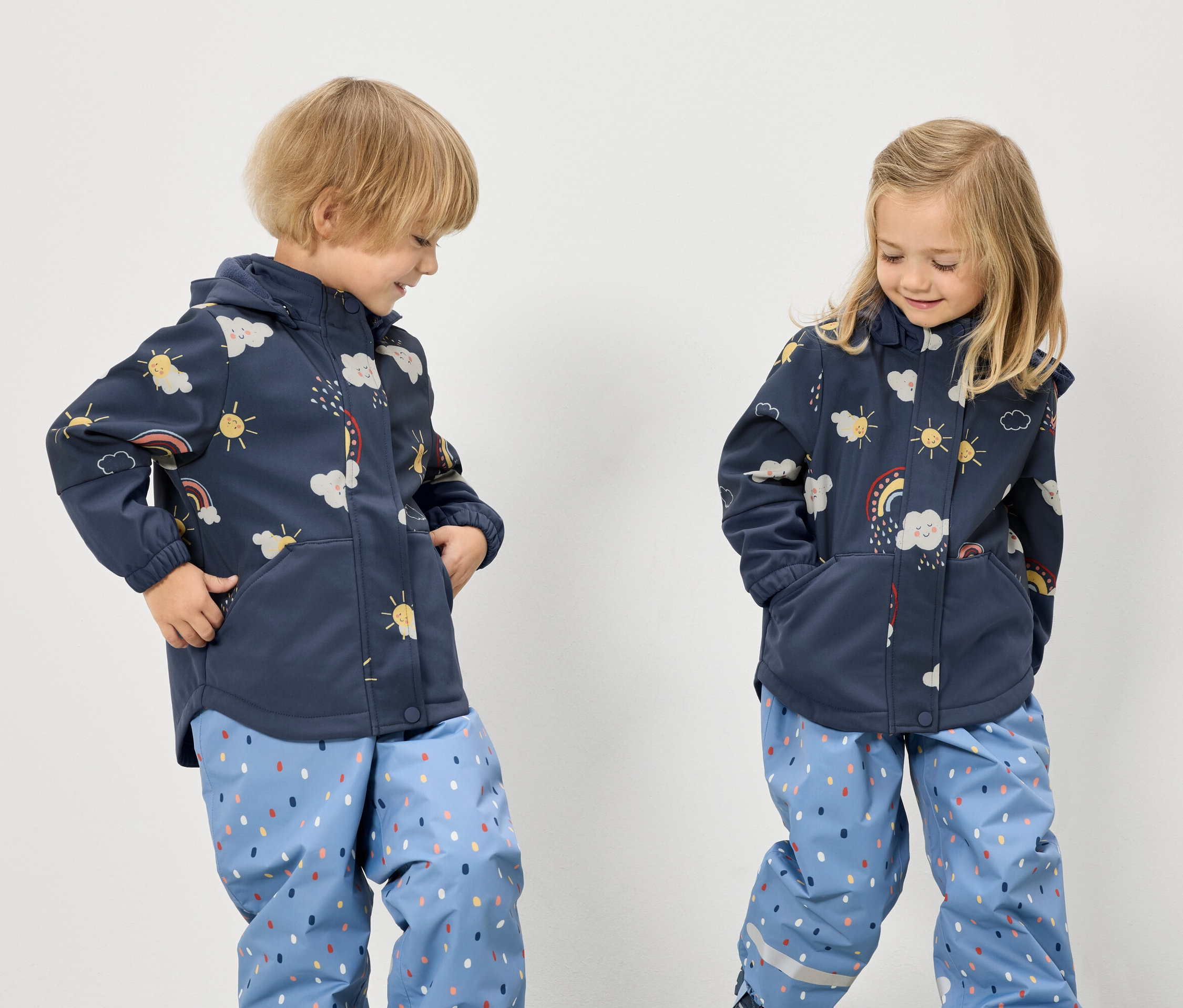 Dve deti v modrých bundách so vzorom dúhy, oblakov a slnka a modrých nohaviciach s bodkovaným vzorom stoja na bielom pozadí.