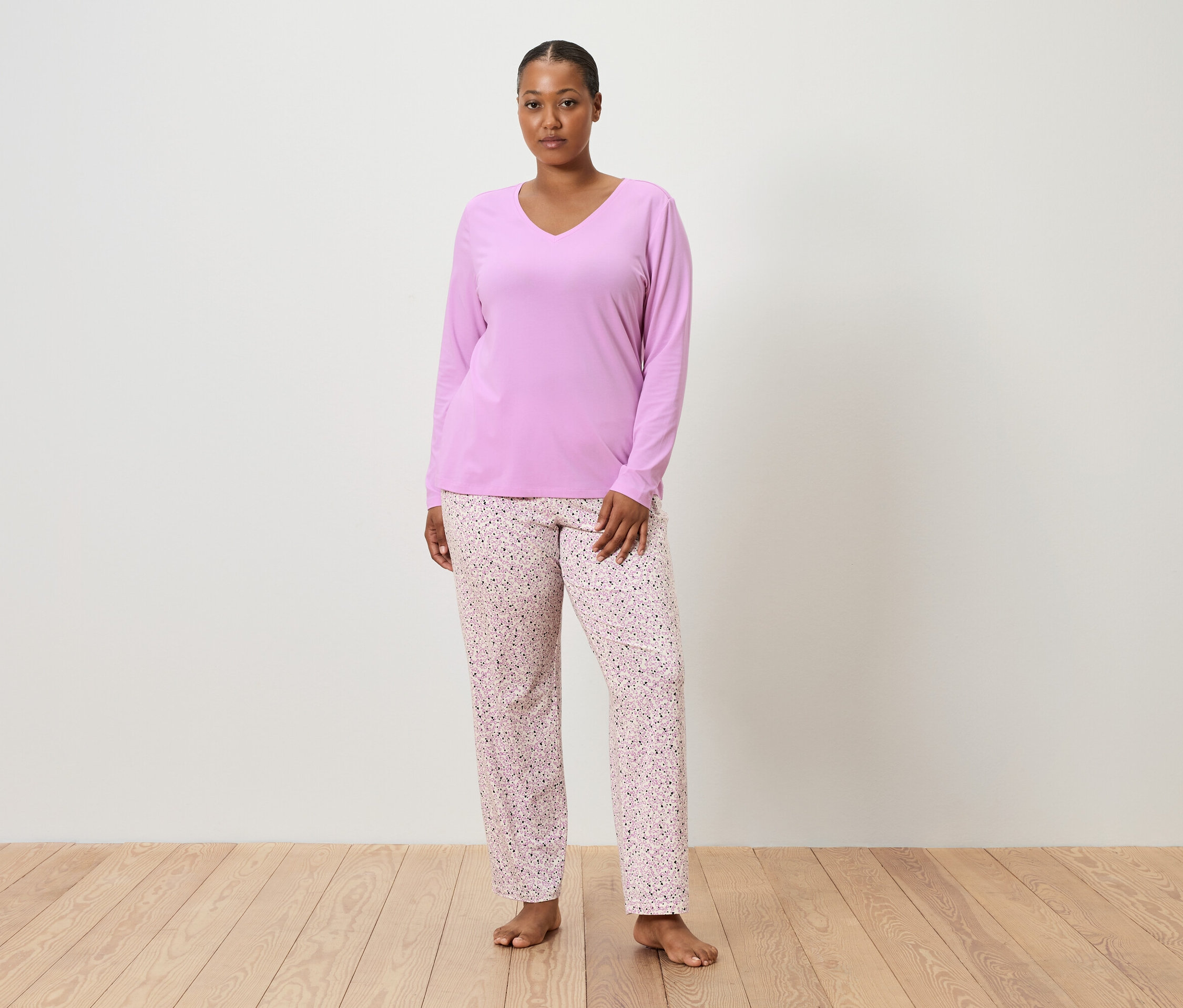 Žena stojí naboso pred bielou stenou a má na sebe fialový pyžamový top a ružové pyžamové nohavice s kvetinovou potlačou.