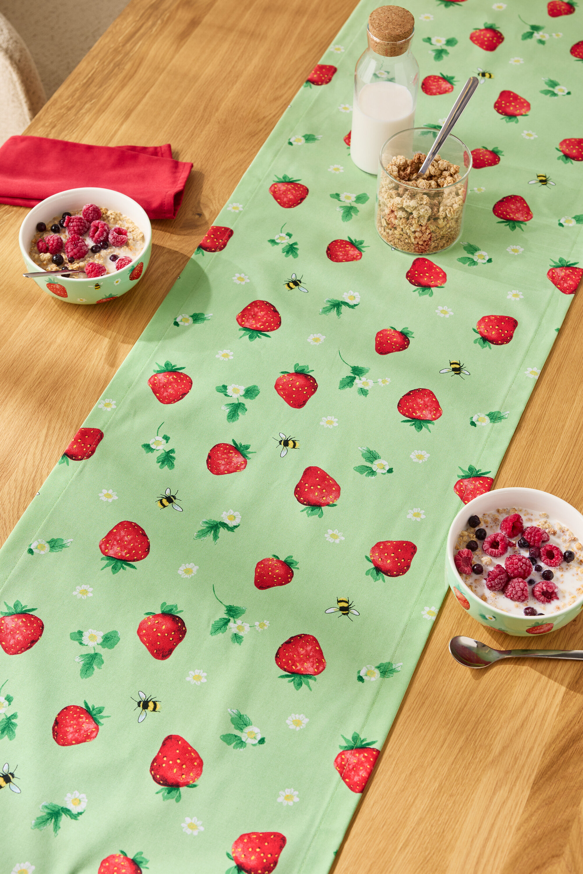 Na drevenom stole leží behúň so vzorom jahôd, na ňom misky s cereáliami, pohár müsli a fľaša mlieka.