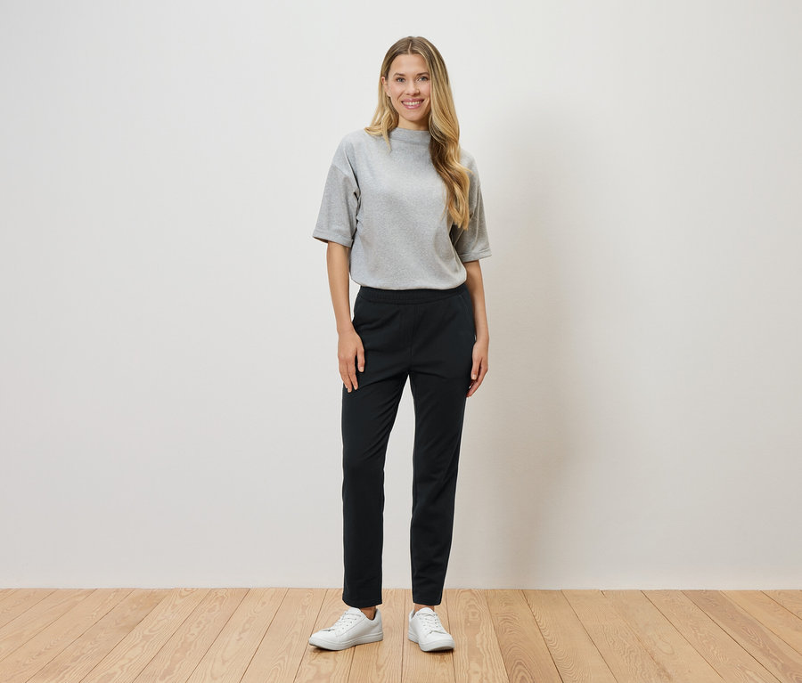 Žena s blond vlasmi stojí pred bielou stenou a má na sebe sivé tričko, čierne nohavice a biele tenisky.