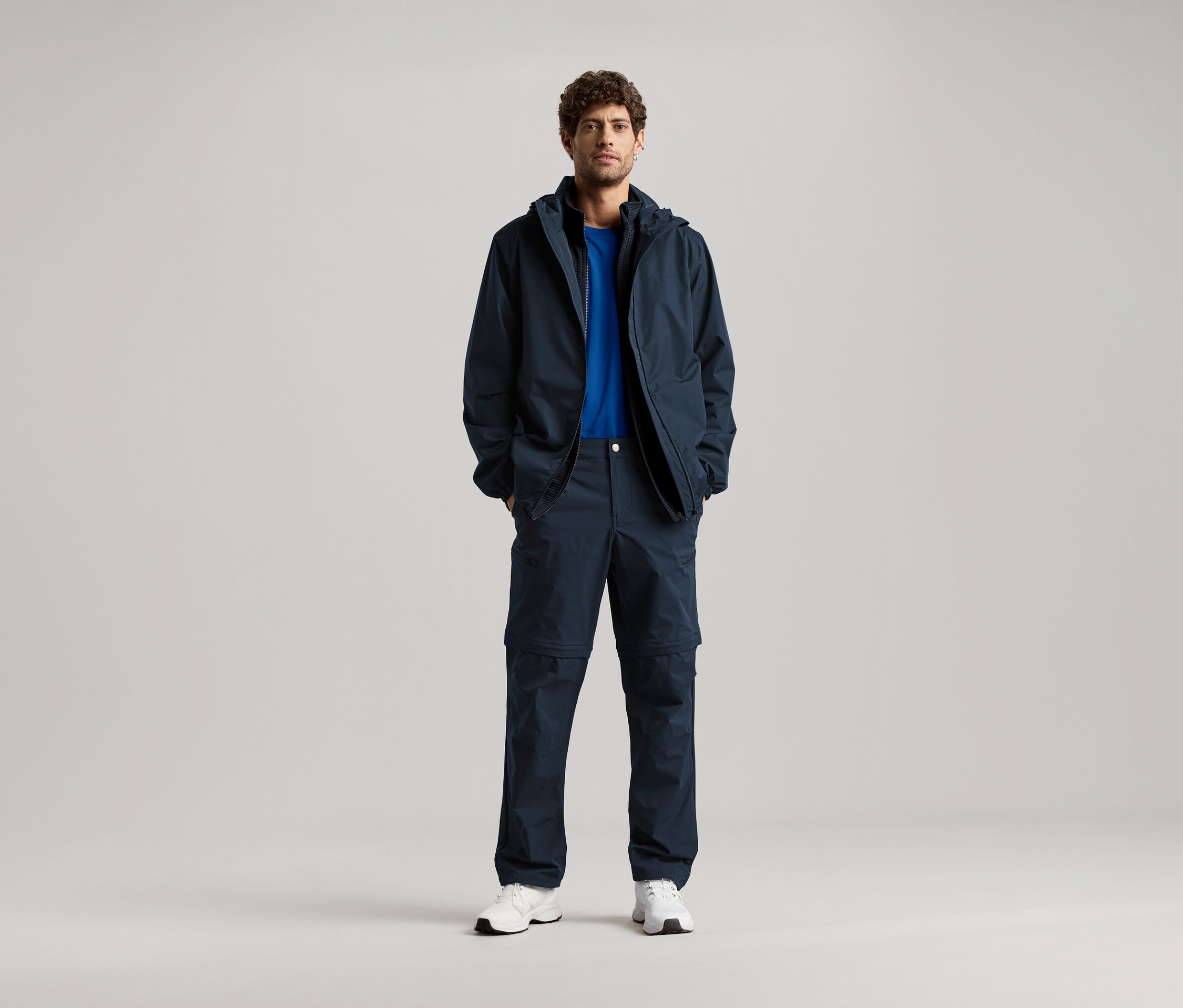 Muž stojí s rukami vo vreckách tmavomodrej bundy a ladiacich nohavíc, má na sebe modré tričko a biele tenisky.