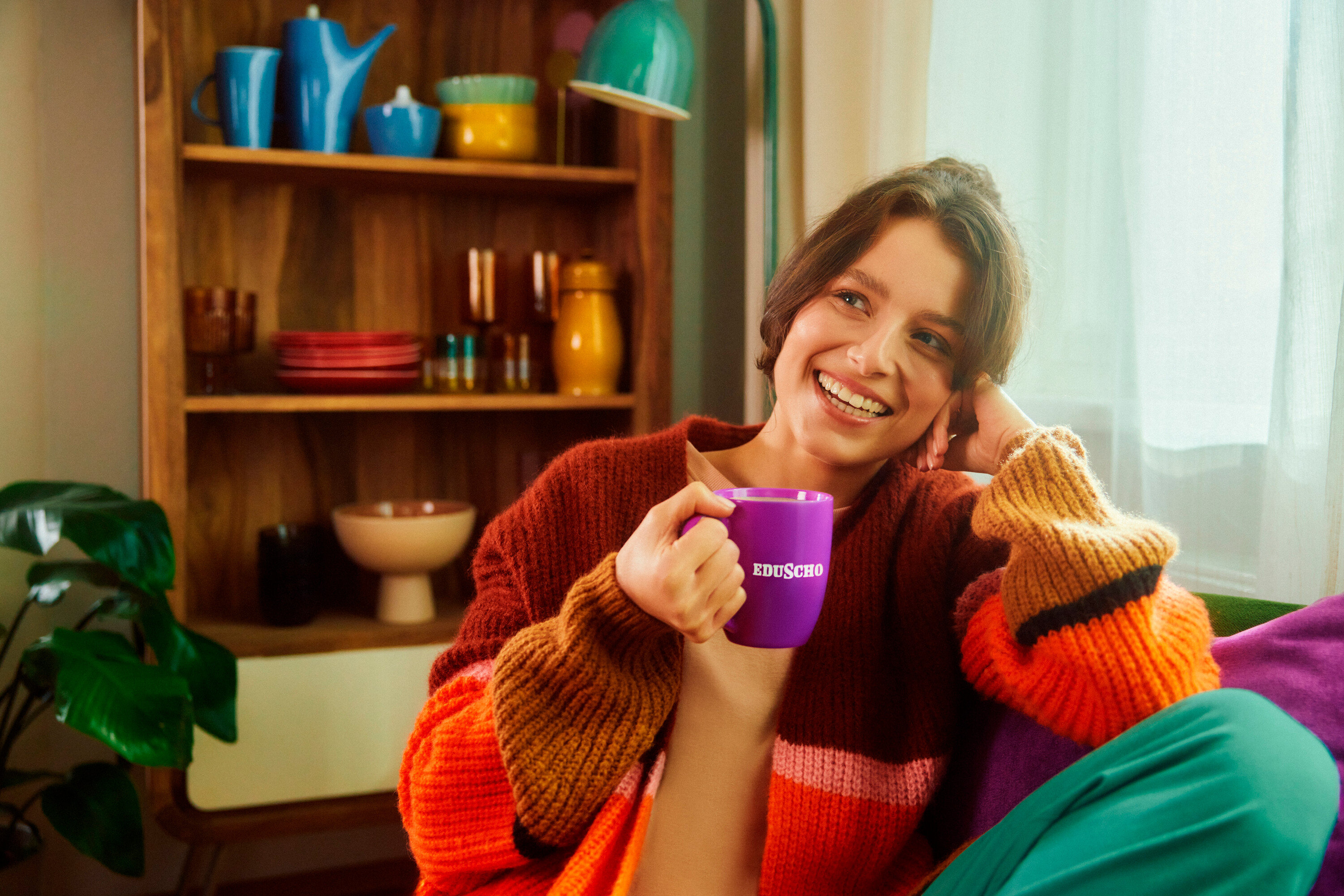 Usmievavá brunetka drží fialový hrnček s nápisom EDUSCHO. V pozadí je polica s riadom.