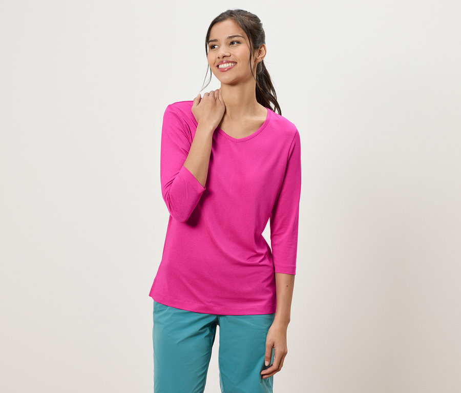 Žena s vrkočom má na sebe ružový top s trojštvrťovými rukávmi a tyrkysové nohavice.