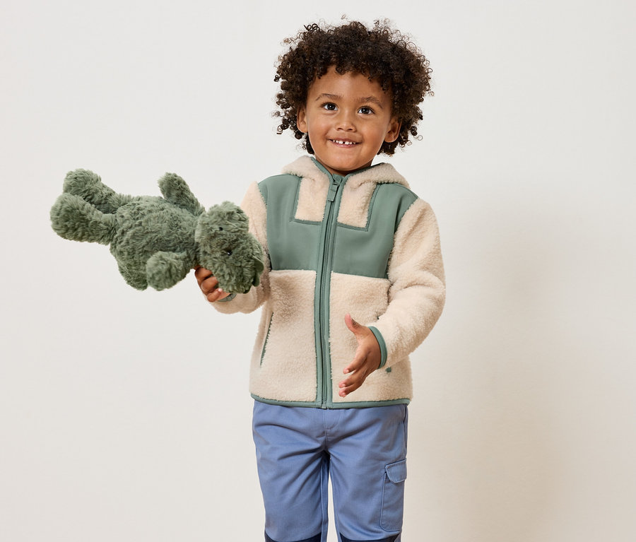 Usmievavý chlapec s kučeravými vlasmi drží zeleného plyšového dinosaura a má na sebe svetlohnedú bundu so zelenými akcentmi a modré nohavice.