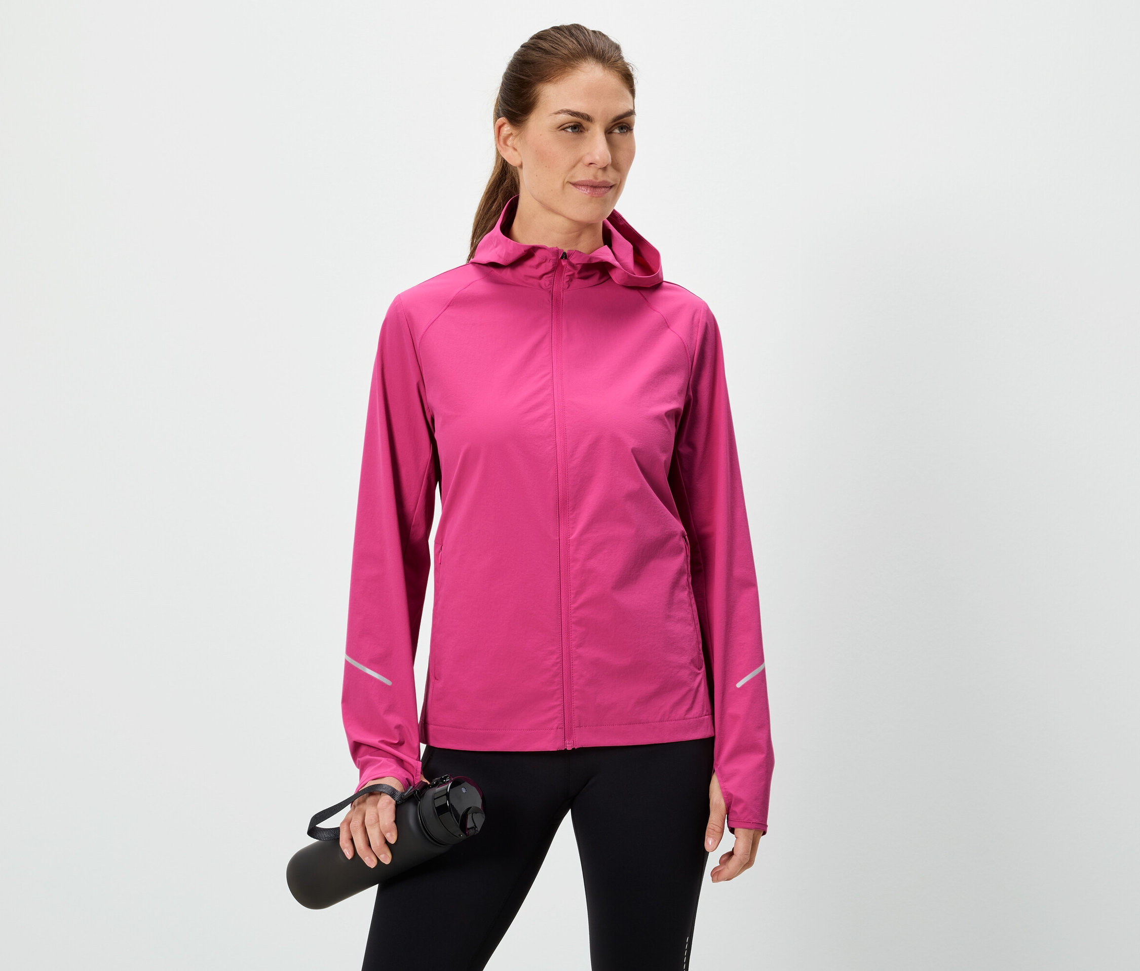 Žena drží čiernu fľašu na vodu, má na sebe ružovú bundu a čierne legíny na bielom pozadí.