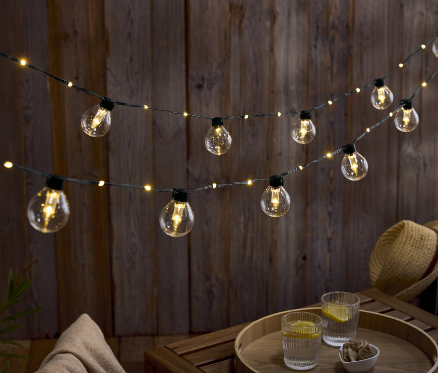 Dva rady žiaroviek visia pred drevenou stenou. Na drevenom stole stoja dva poháre vody s citrónom a miska orechov.