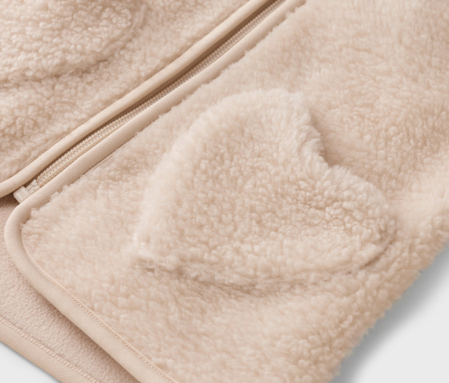 Detailný záber na krémovú teddy bundu so zipsom a vreckom v tvare srdca.