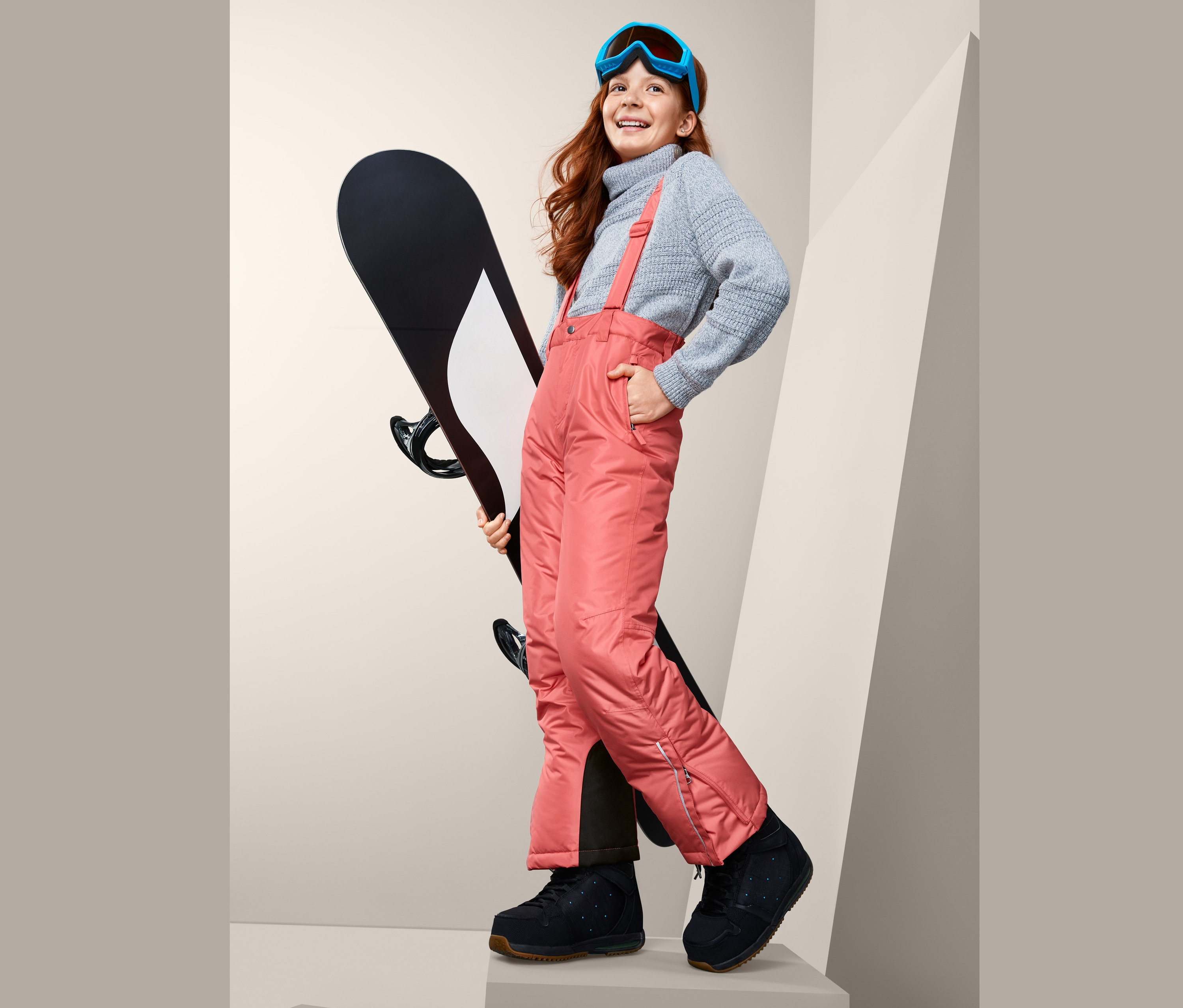 Usmievajúce sa dievča drží snowboard a má na sebe lyžiarske nohavice, sveter a lyžiarske okuliare.