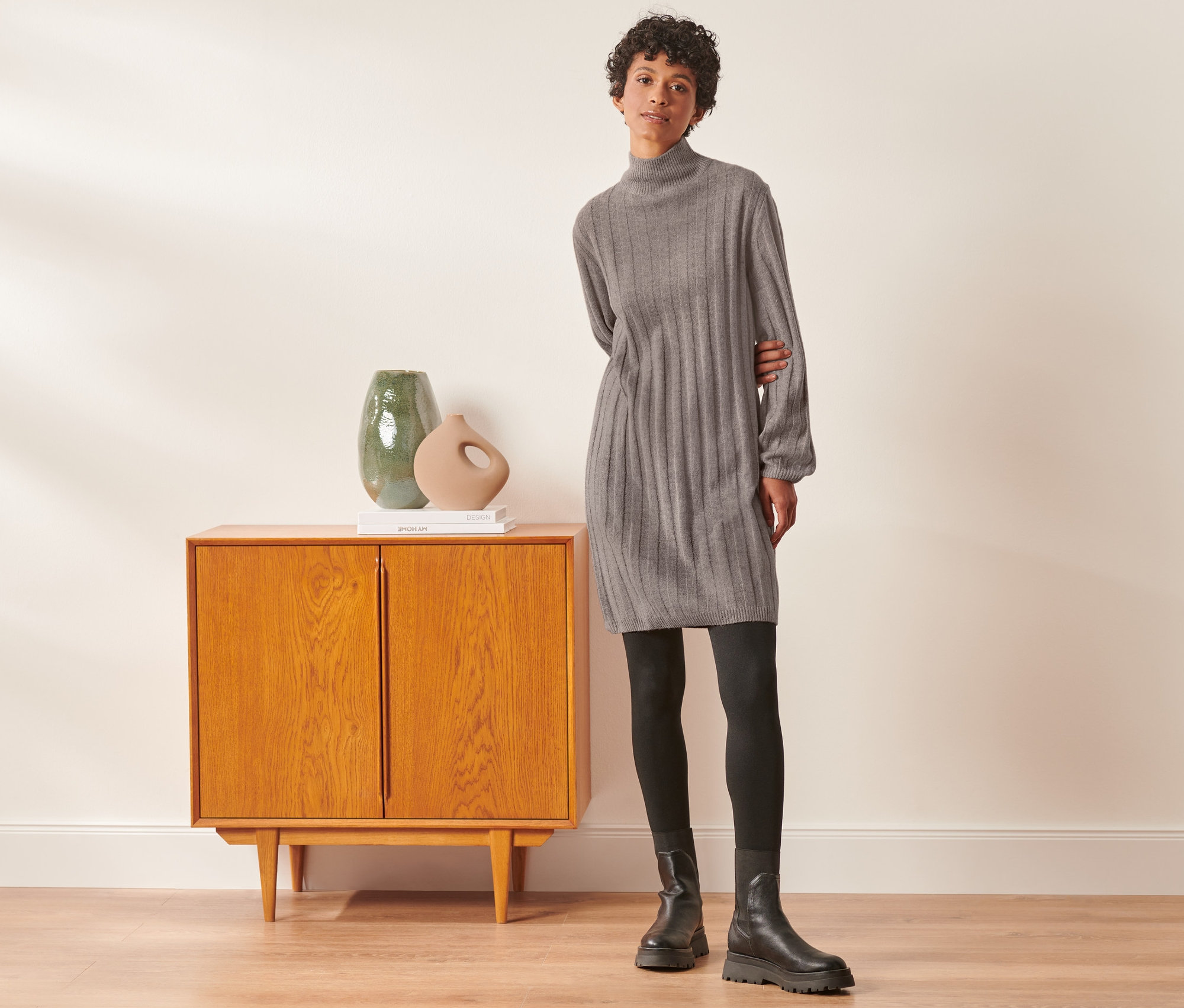 Žena pózuje vedľa skrine a má na sebe rebrované pletené šaty so stojačikom, čierne legíny a topánky.
