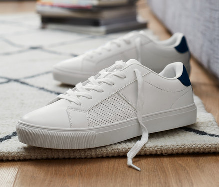 Dvoje topánky sneaker ležia na podlahe vedľa šedej pohovky.