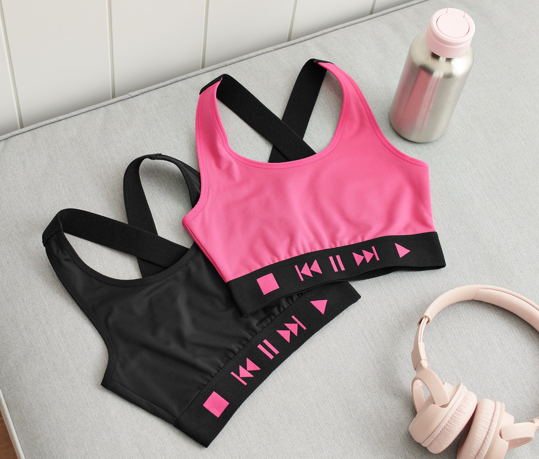 Dve detské športové podprsenky, jedna ružová a jedna čierna, ležia na sivej pohovke vedľa slúchadiel, fľaše s vodou a topánok.