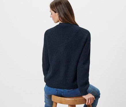 Žena sedí na drevenej stoličke a má na sebe tmavomodrý kardigán z hrubej pleteniny.