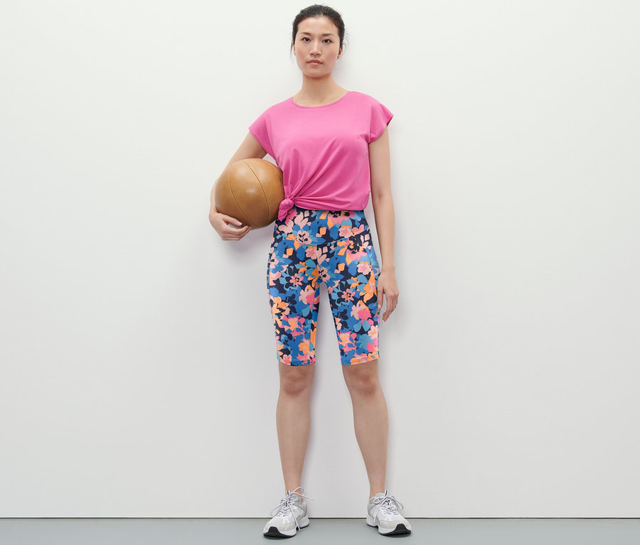 Žena drží medicinbal a má na sebe športové cyklistické šortky s celoplošnou potlačou, tričko a tenisky.