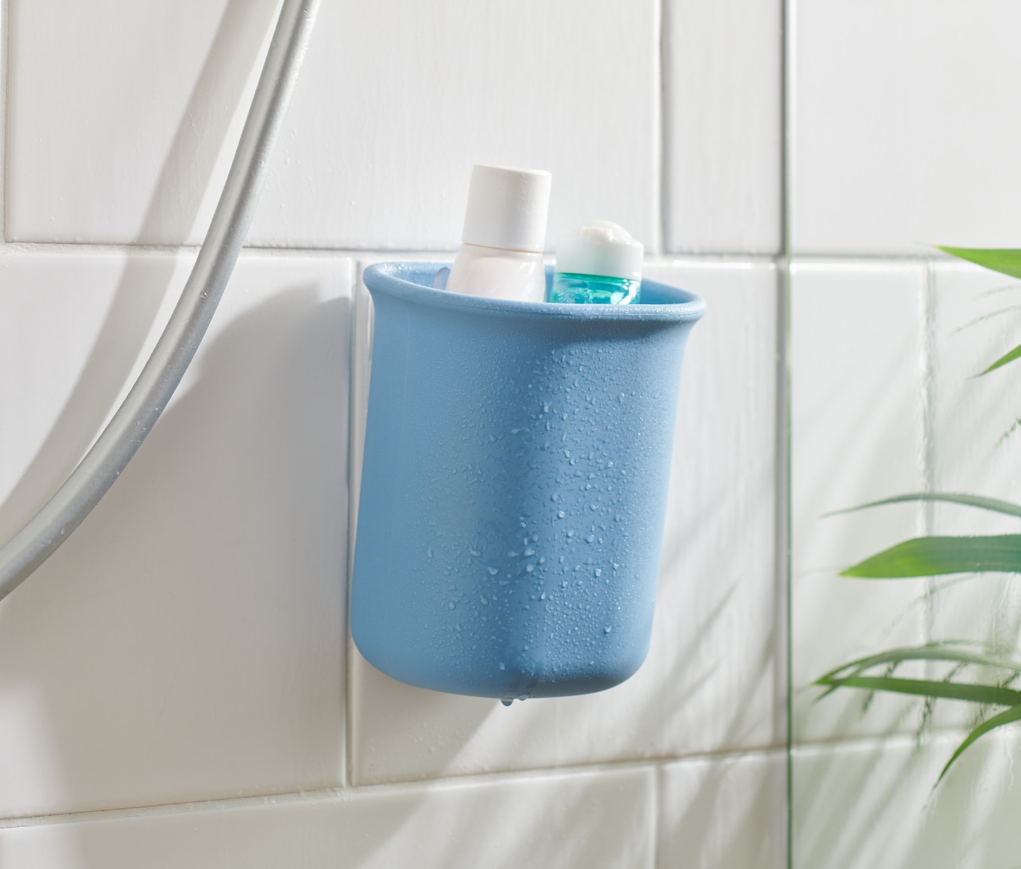 Modrý kúpeľňový organizér s bielym a modrým produktom vo vnútri, pripevnený na bielych dlaždiciach vedľa sprchovej hadice.