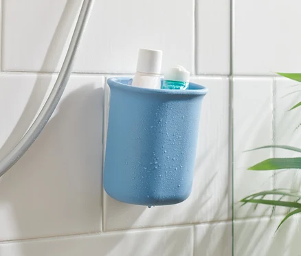 Modrý kúpeľňový organizér s bielym a modrým produktom vo vnútri, pripevnený na bielych dlaždiciach vedľa sprchovej hadice.