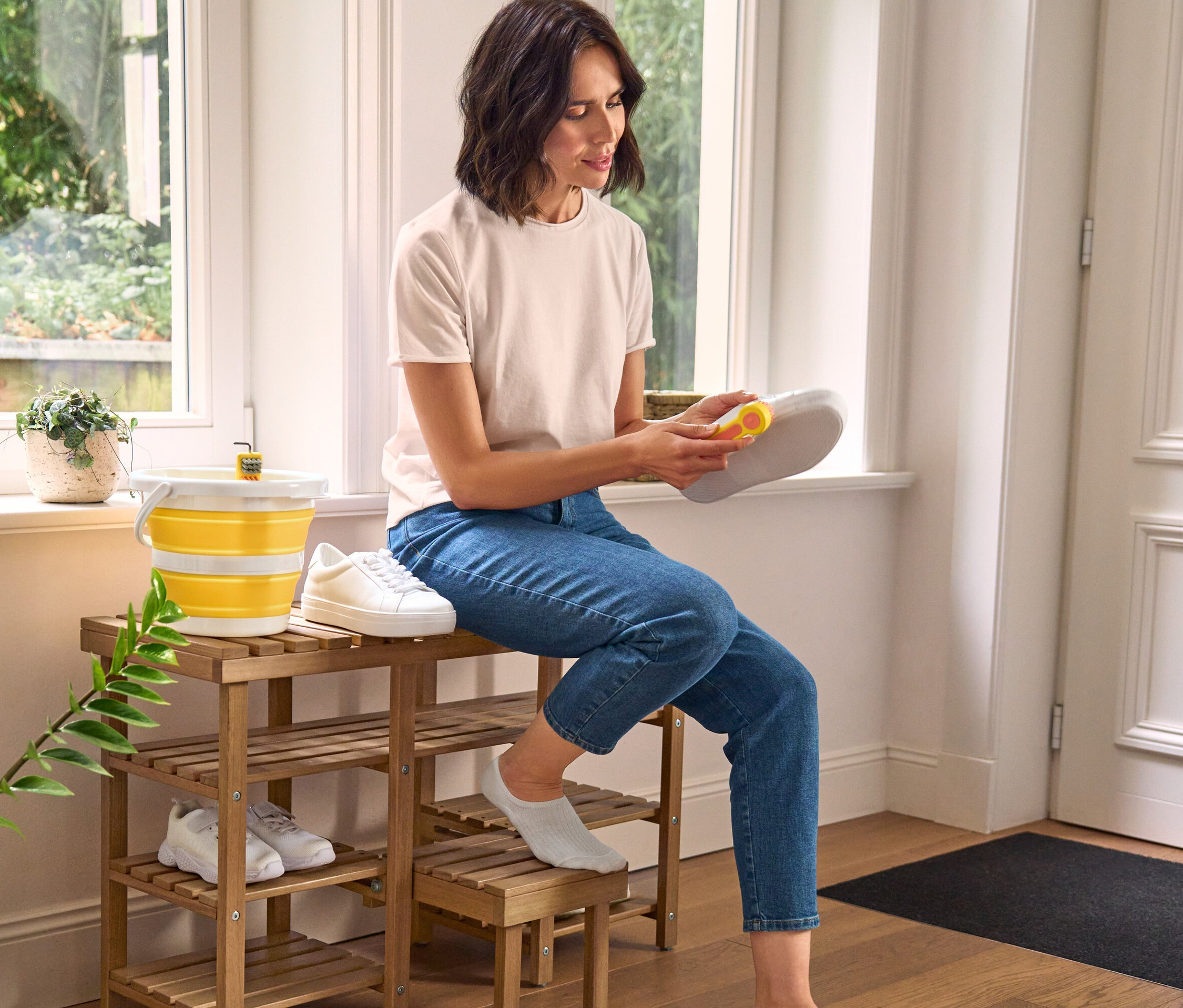 Žena čistí topánku kefou na čistenie obuvi 3 v 1, sedí na vysokej sedacej lavici na topánky. Vedľa nej stojí skladacie vedro a kefa na topánky.