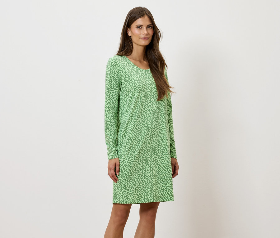 Žena stojí a pózuje v zelených vzorovaných šatách s dlhým rukávom.