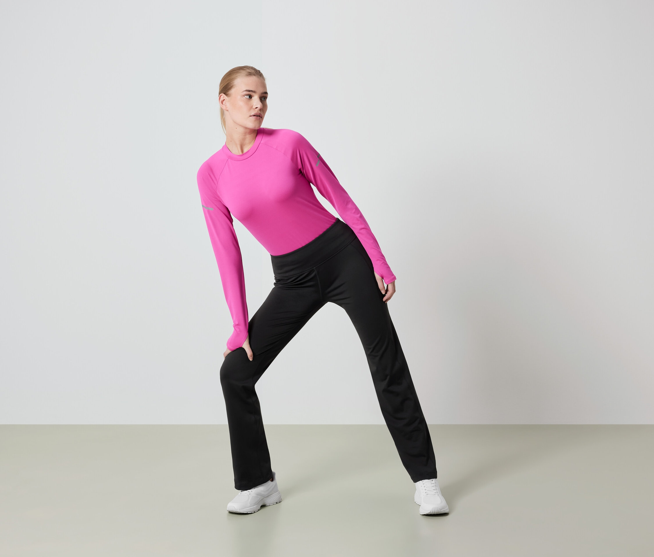 Žena so svetlými vlasmi sa pretahuje v ružovom tope s dlhým rukávom a čiernych nohaviciach.