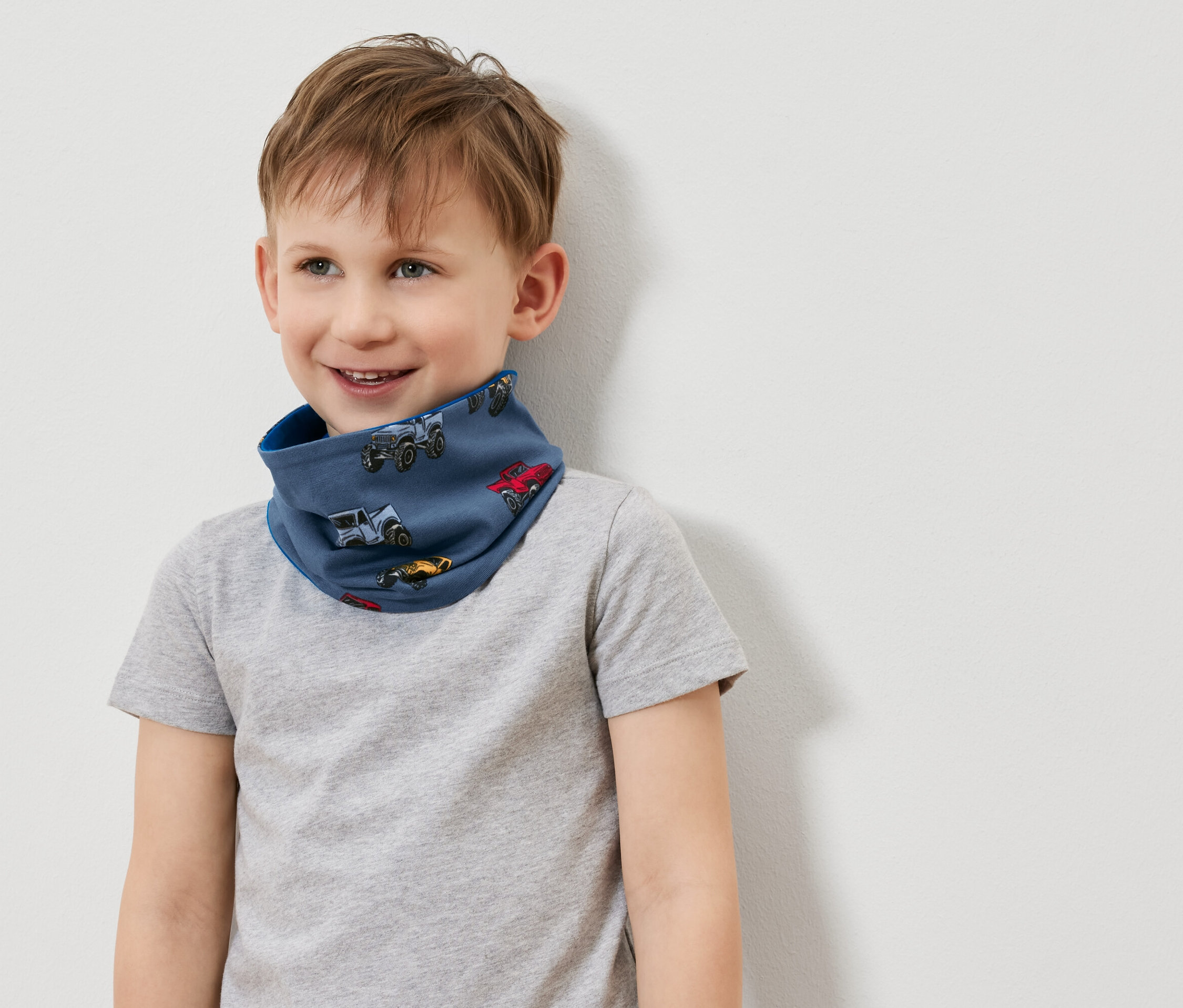 Usmievavý chlapec nosí modrý nákrčník s potlačou autíčok a sivé tričko.