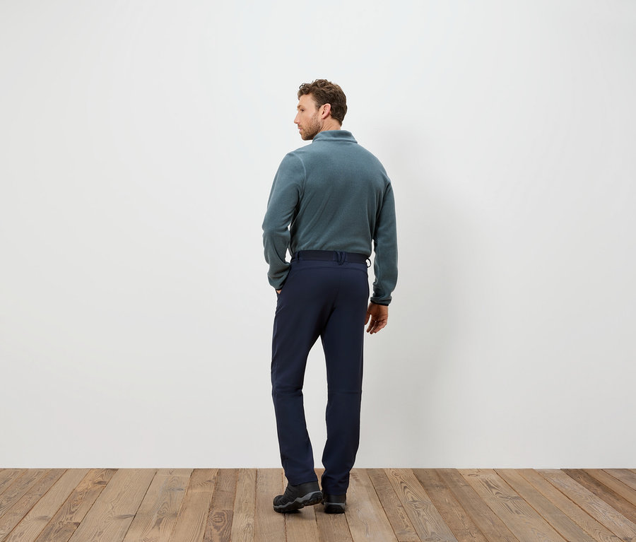 Muž v modrých nohaviciach a flísovej mikine stojí chrbtom ku kamere na drevenej podlahe.
