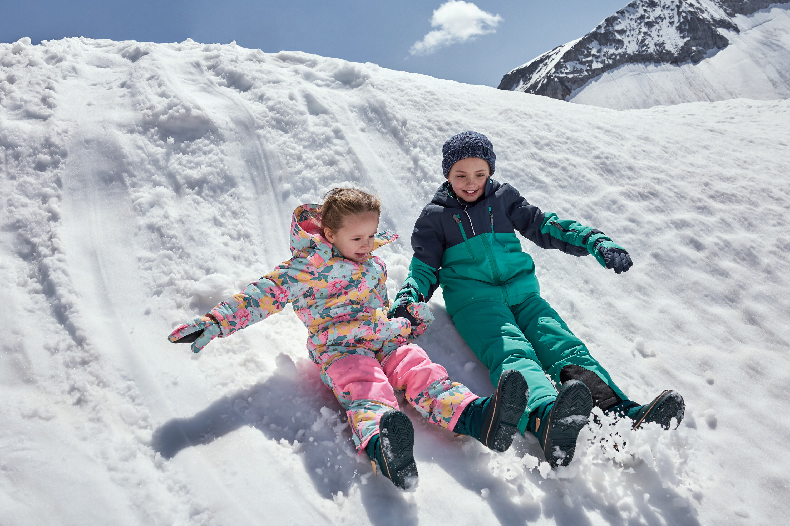 Deti sa spúšťajú po zasneženom kopci v farebných zimných kombinézach.
