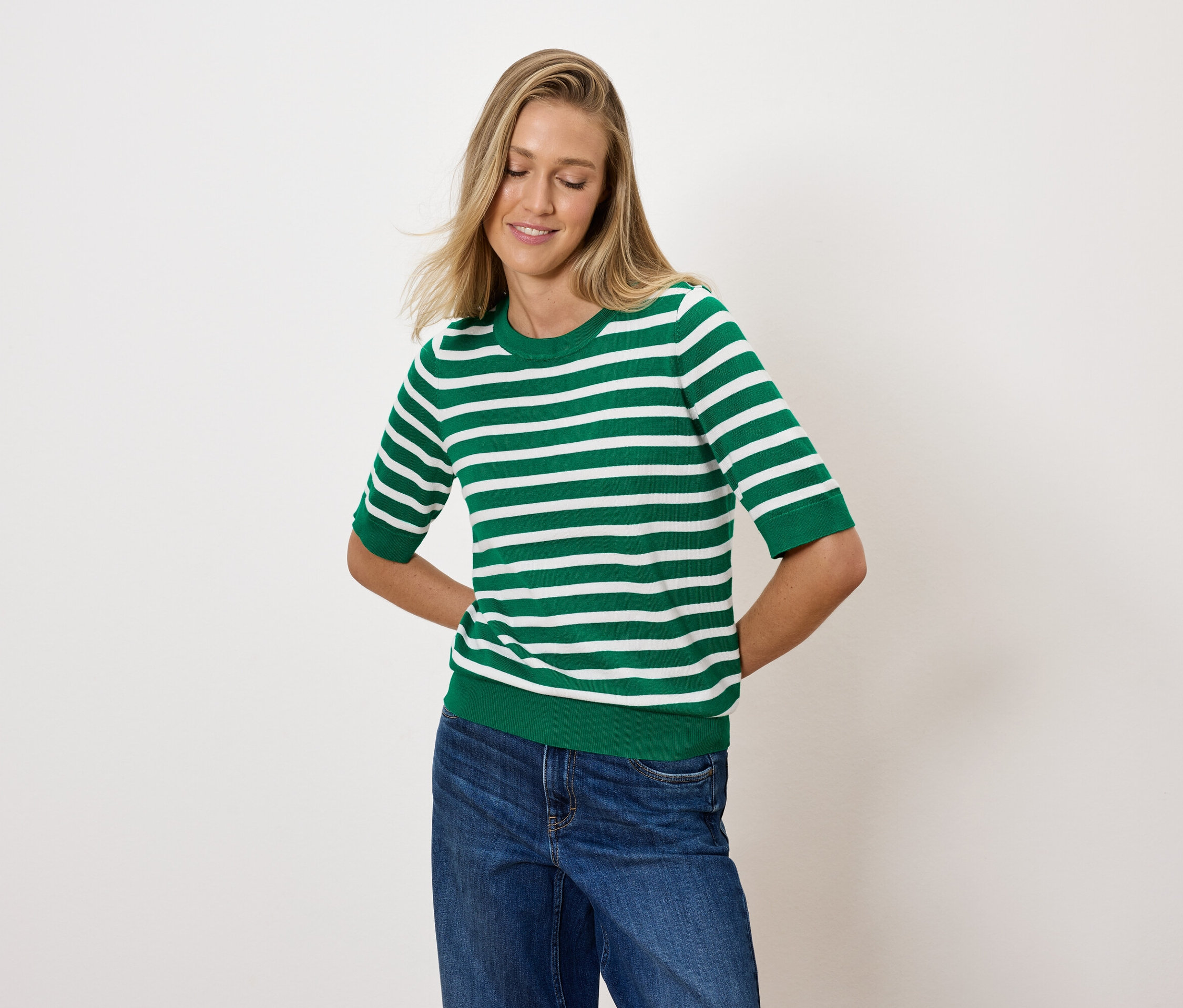 Žena s blond vlasmi pózuje v zeleno-bielom pruhovanom tope a modrých džínsoch.