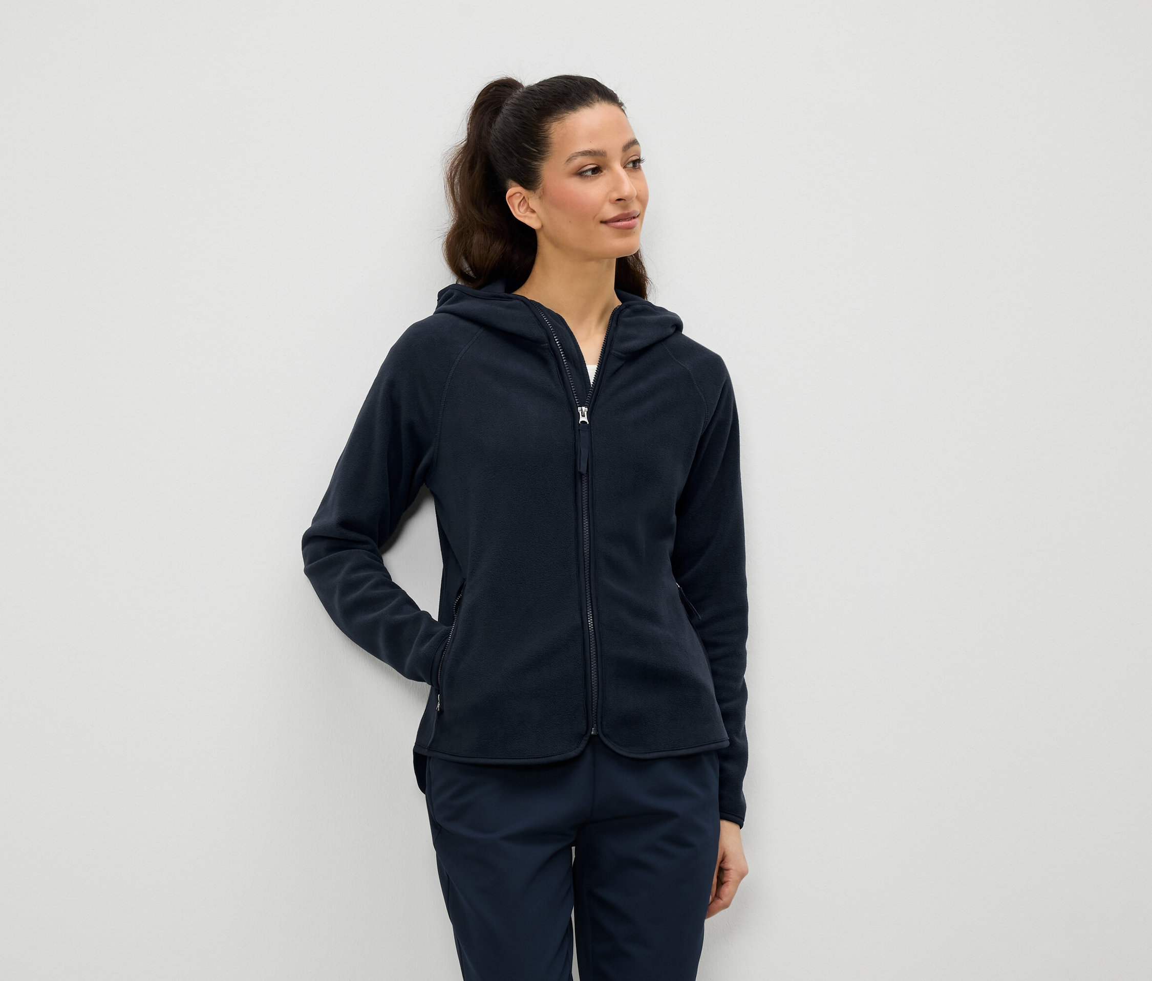 Žena má na sebe tmavomodrú fleecovú bundu s kapucňou a zipsom a tmavomodré nohavice.