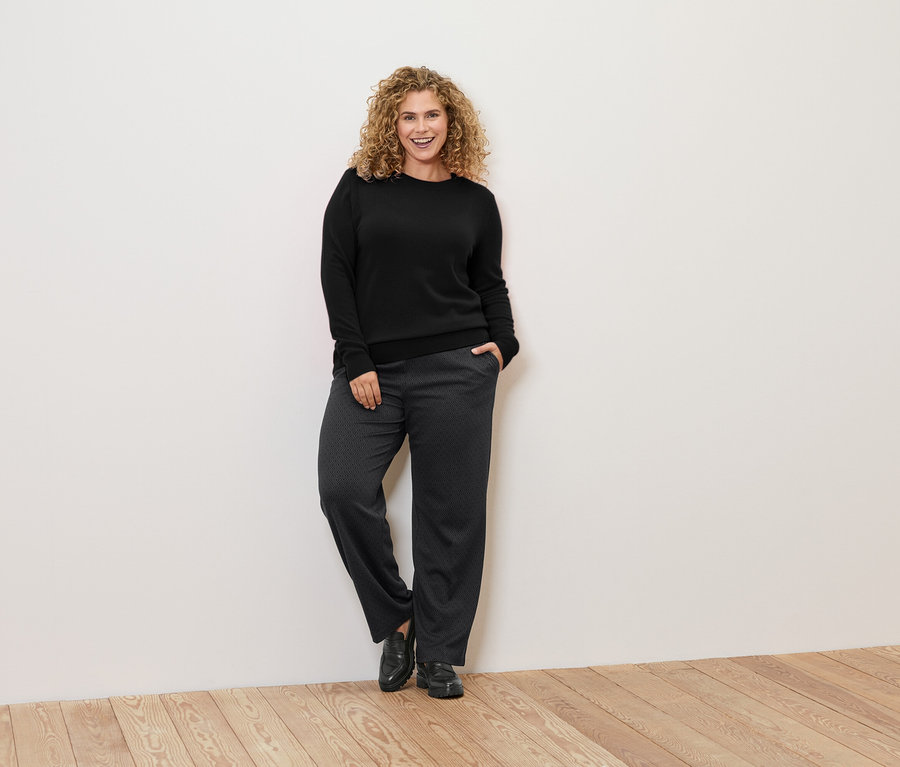 Žena s kučeravými vlasmi sa opiera o stenu, má na sebe čierny sveter a vzorované nohavice.