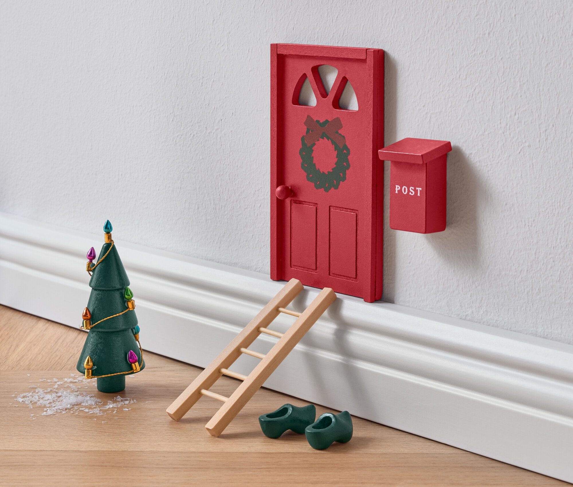 Miniatúrne červené dvere zdobené vencom s rebríkom, zelenými drevákmi a vianočným stromčekom.