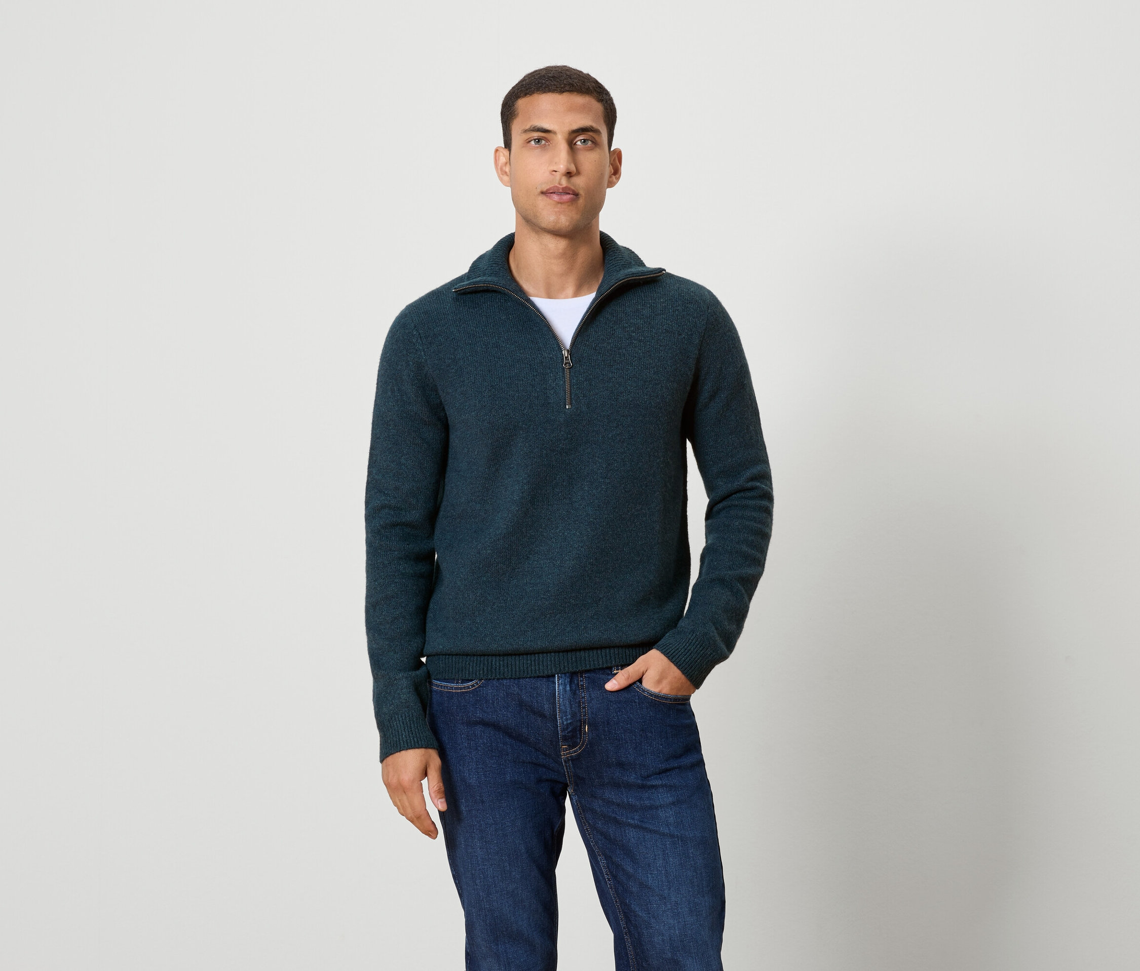 Muž pózuje na pozadí a má na sebe tmavomodrý sveter na zips a džínsy.