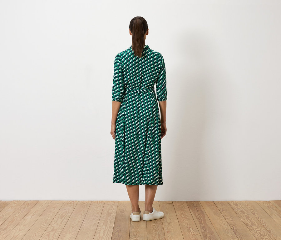 Žena stojí chrbtom k fotoaparátu a má na sebe zelené šaty s bielym vzorom a biele tenisky.