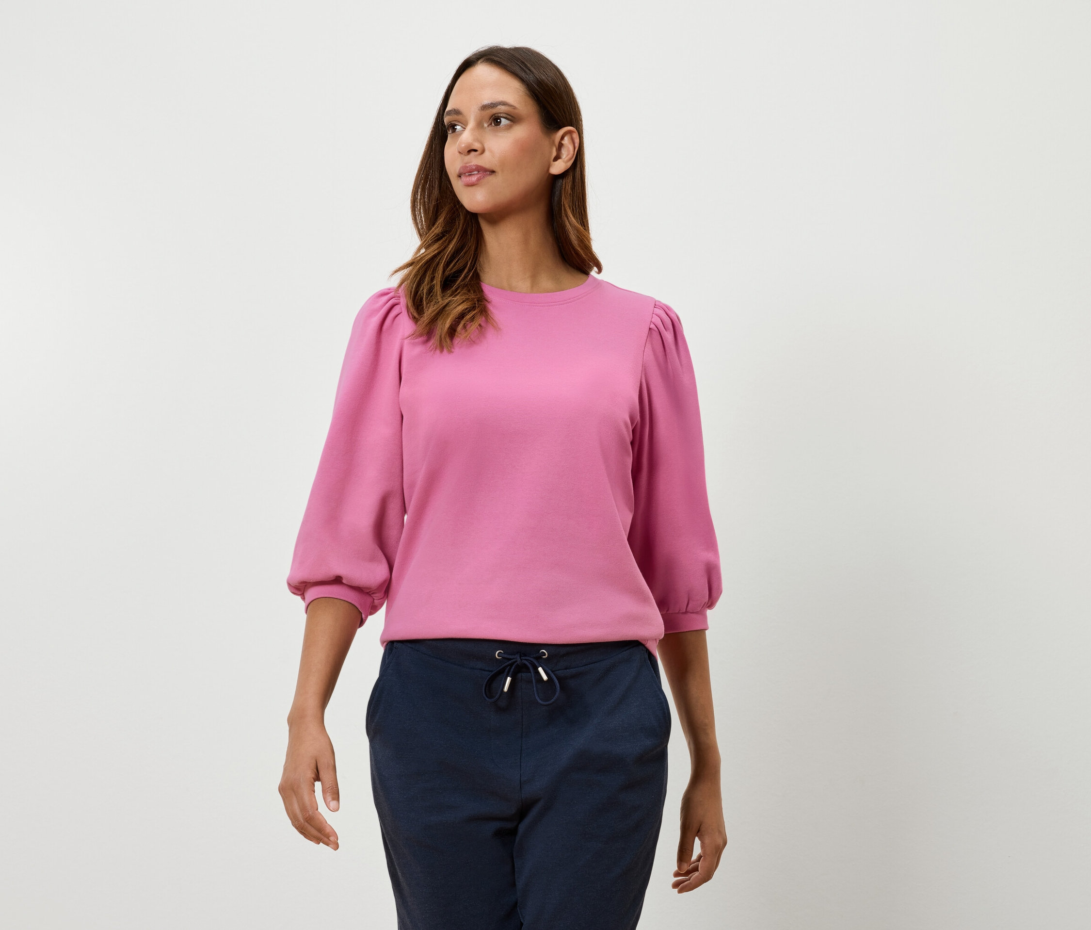 Žena má na sebe ružovú mikinu s trojštvrťovými balónovými rukávmi a tmavomodré nohavice.