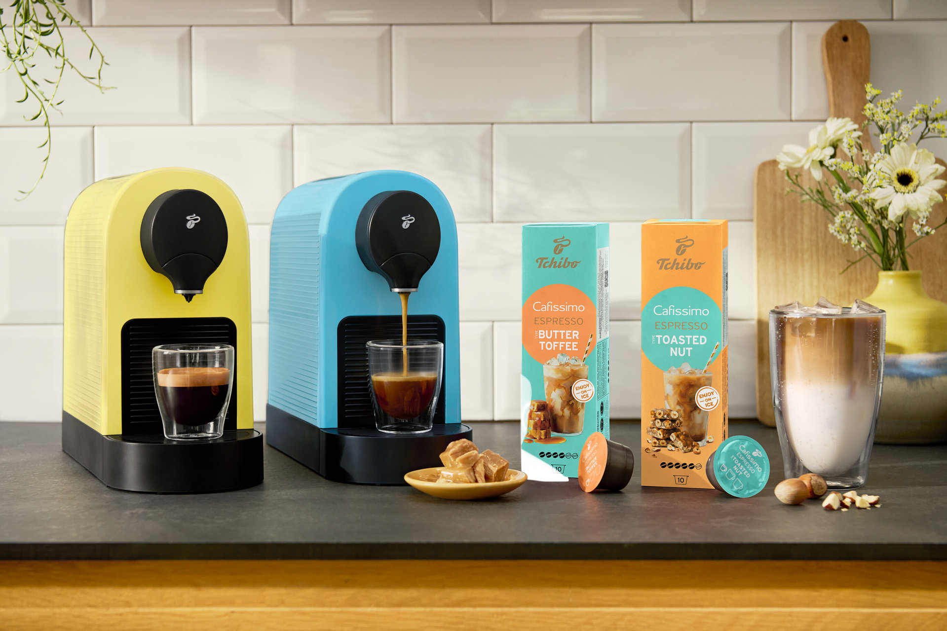 Dva kávovary Cafissimo pure+ v žltej a modrej farbe pripravujú espresso. Vidieť sú aj kapsuly Cafissimo Flavoured Espresso – Toasted Nut a Buttertoffee.