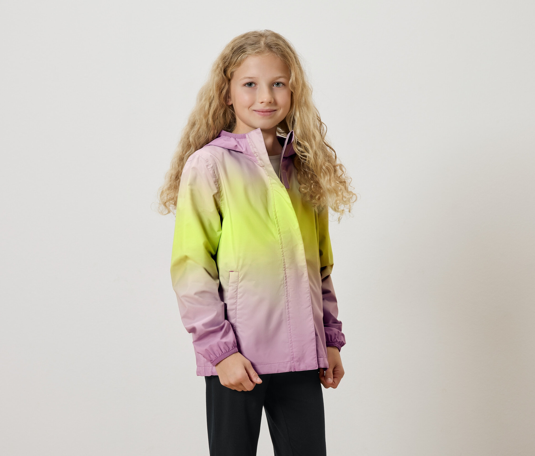 Dievča s dlhými blond kučerami má na sebe bundu s kapucňou s farebným prechodom zo žltej do fialovej.