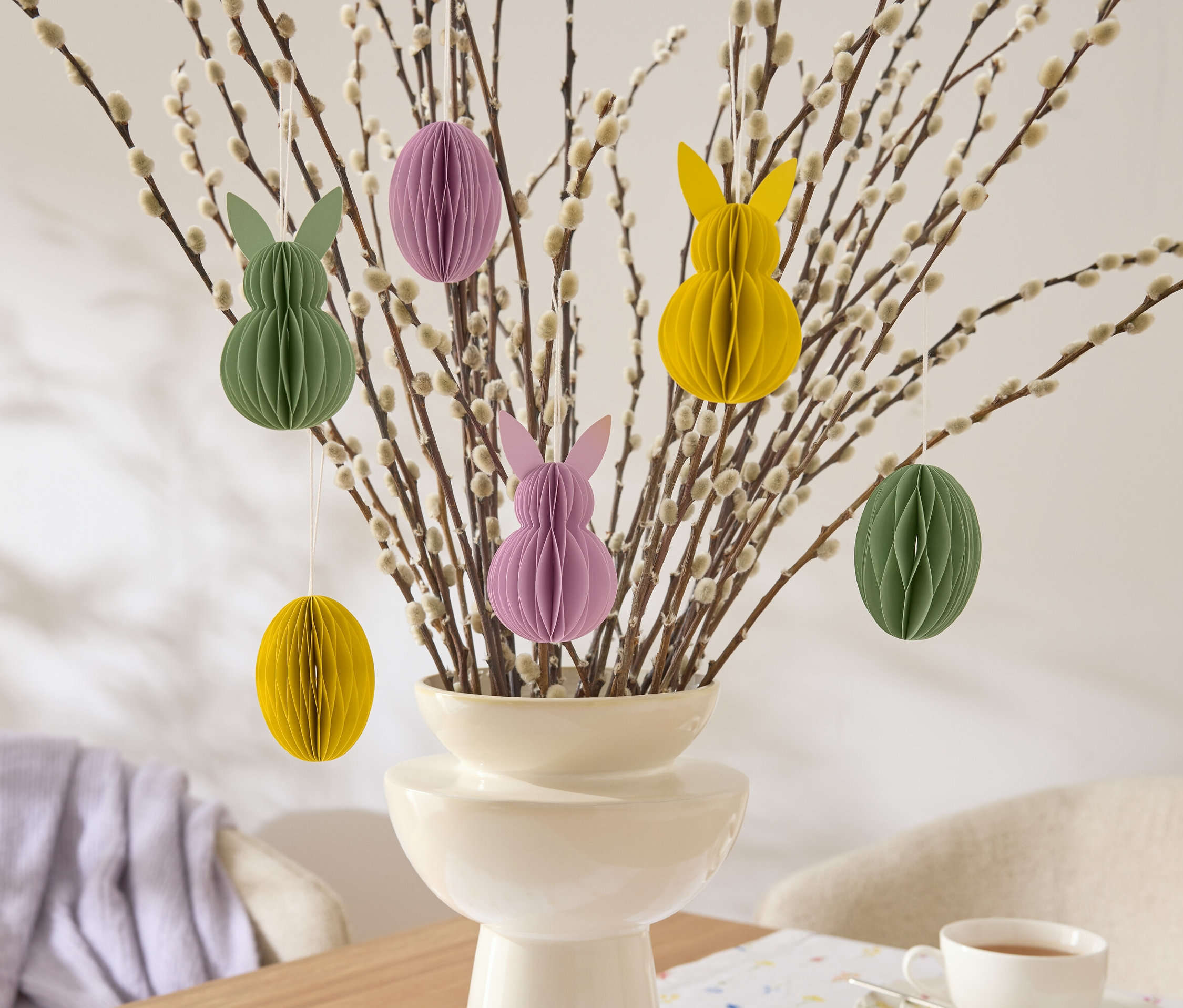 Dekoratívne veľkonočné vetvičky s visiacimi papierovými ozdobami v tvare zajačikov a vajíčok v žltej, zelenej a fialovej farbe vo váze.